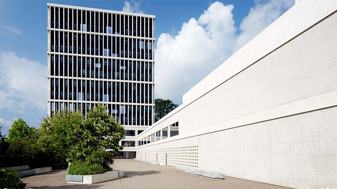 Photo shows the Federal Administrative Court building in St. Gallen. The sky is blue with white clouds and the building is grey/white. In front of the building, you can see trees and shrubs. The building itself is a modern high-rise building with many large windows.