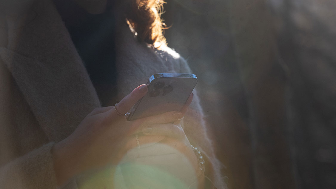 Una signora dai capelli scuri indossa abiti autunnali e in mano ha uno smartphone. Nell'immagine compare il sole.