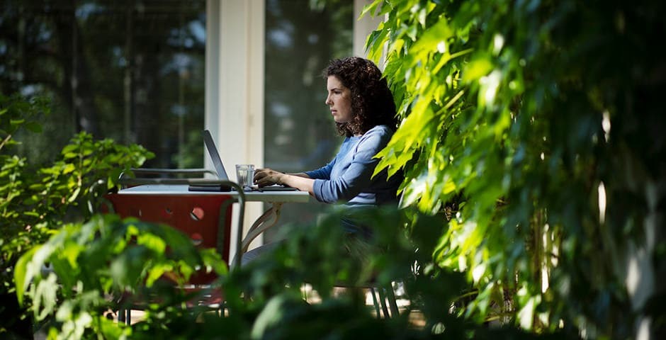 Eine Frau sitzt draussen im Garten an einem Tisch, vor ihr der Laptop und ein Glas Wasser.